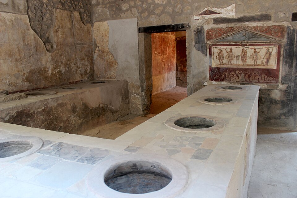 The stone counter of a thermopolium in Pompeii, a Roman fast-food shop, with large round ceramic jars (dolia) set into the masonry of the L-shaped serving counter where hot food and drink were kept