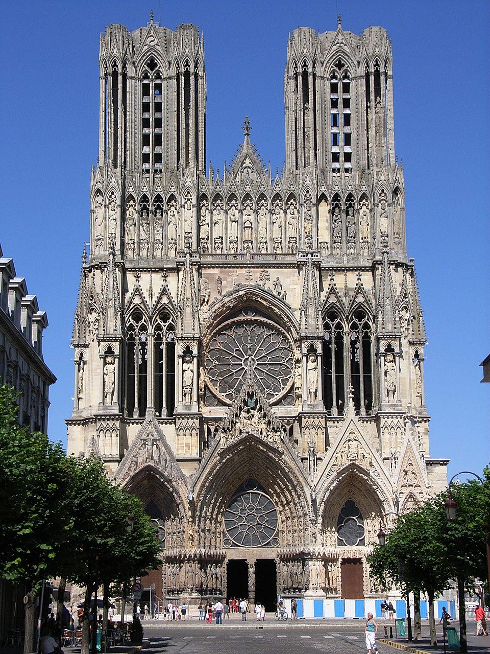 The soaring Gothic interior of Reims Cathedral, with immensely tall stone columns rising to a vaulted ceiling far overhead and light pouring through stained-glass windows at the far end