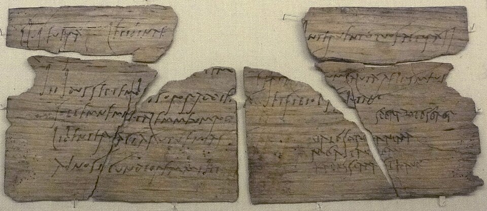 A thin postcard-sized wooden writing tablet from Vindolanda, covered in faint lines of cursive Latin written in carbon ink, with two visibly different handwriting styles &mdash; the scribe's elegant hand and Claudia Severa's own less formal closing