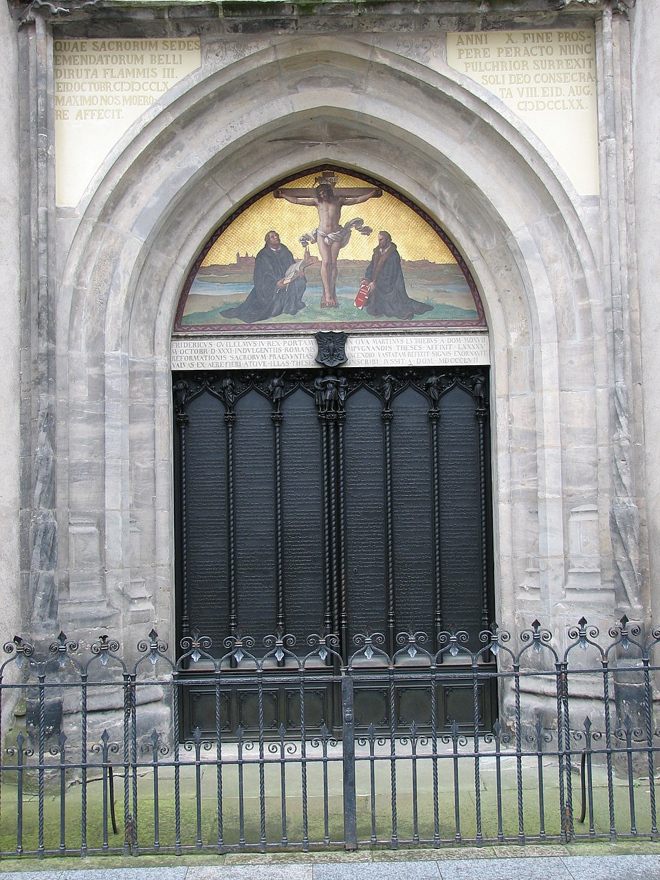 A close view of the bronze double-doors of the Castle Church in Wittenberg, with the full Latin text of Luther&rsquo;s Ninety-Five Theses cast into the metal in many small lines, framed by a stone Gothic arch above